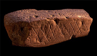 Kus červeného okru s vrypy, které udělal obyvatel jeskyně Blombos před přibližně 70.000 lety (Ch. S. Henshilwood, https://creativecommons.org/licenses/by-sa/3.0/deed.en, CC BY-SA 3.0, via Wikimedia Commons).